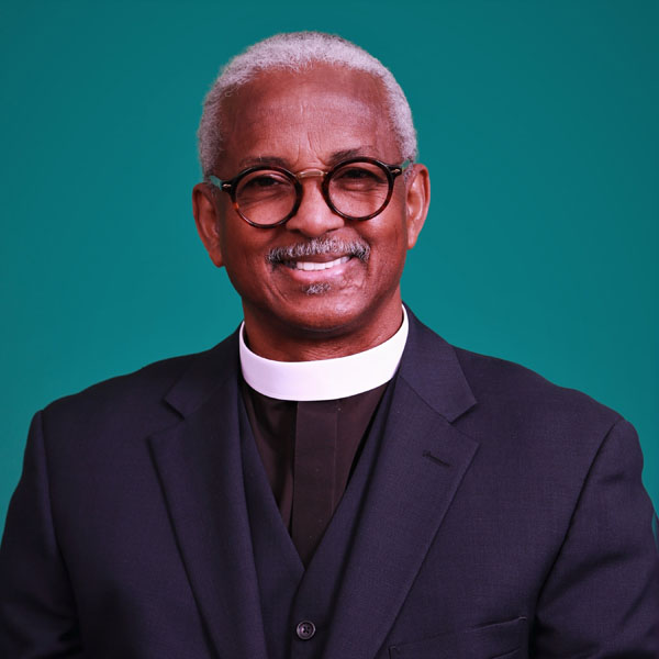 Reverend Anthony Thompson wearing a black clerical suit and white collar, smiling in front of a teal background.