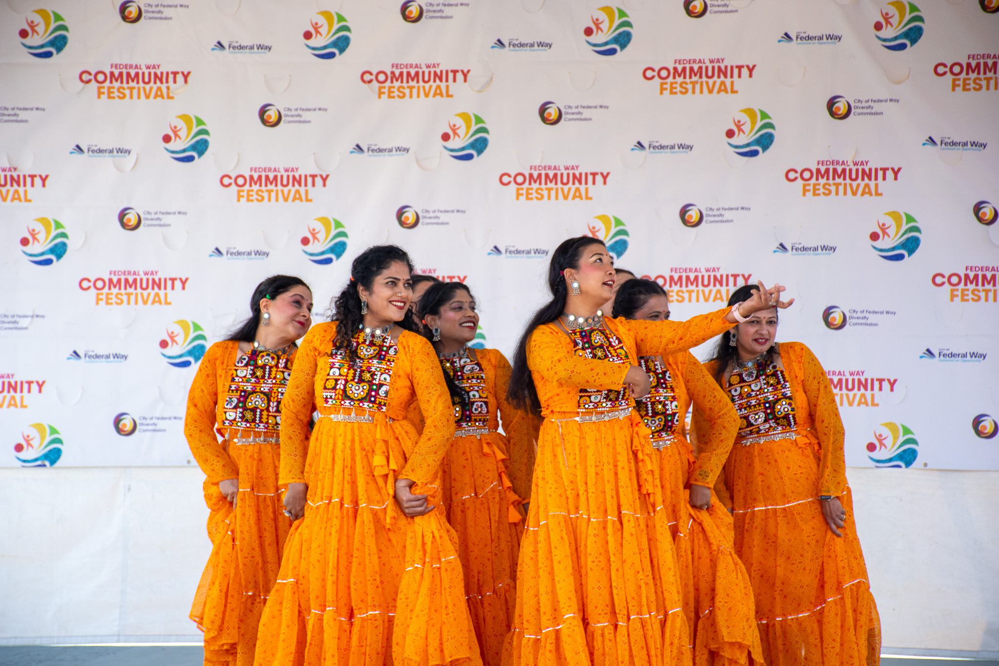 Five women in bright orange traditional dresses pose together at the Federal Way Community Festival, smiling and gesturing on a stage in front of a Community Festival backdrop.