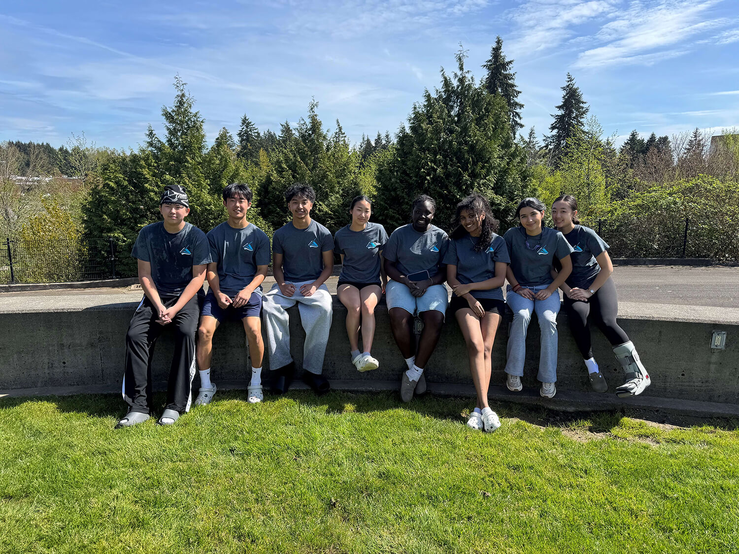 Group of eight young people wearing matching Federal Way Youth Commission dark blue T-shirts sit on a low concrete wall outdoors on a sunny day, with green grass in the foreground and tall evergreen trees behind them. The group faces the camera, some smiling, under a blue sky with light clouds.